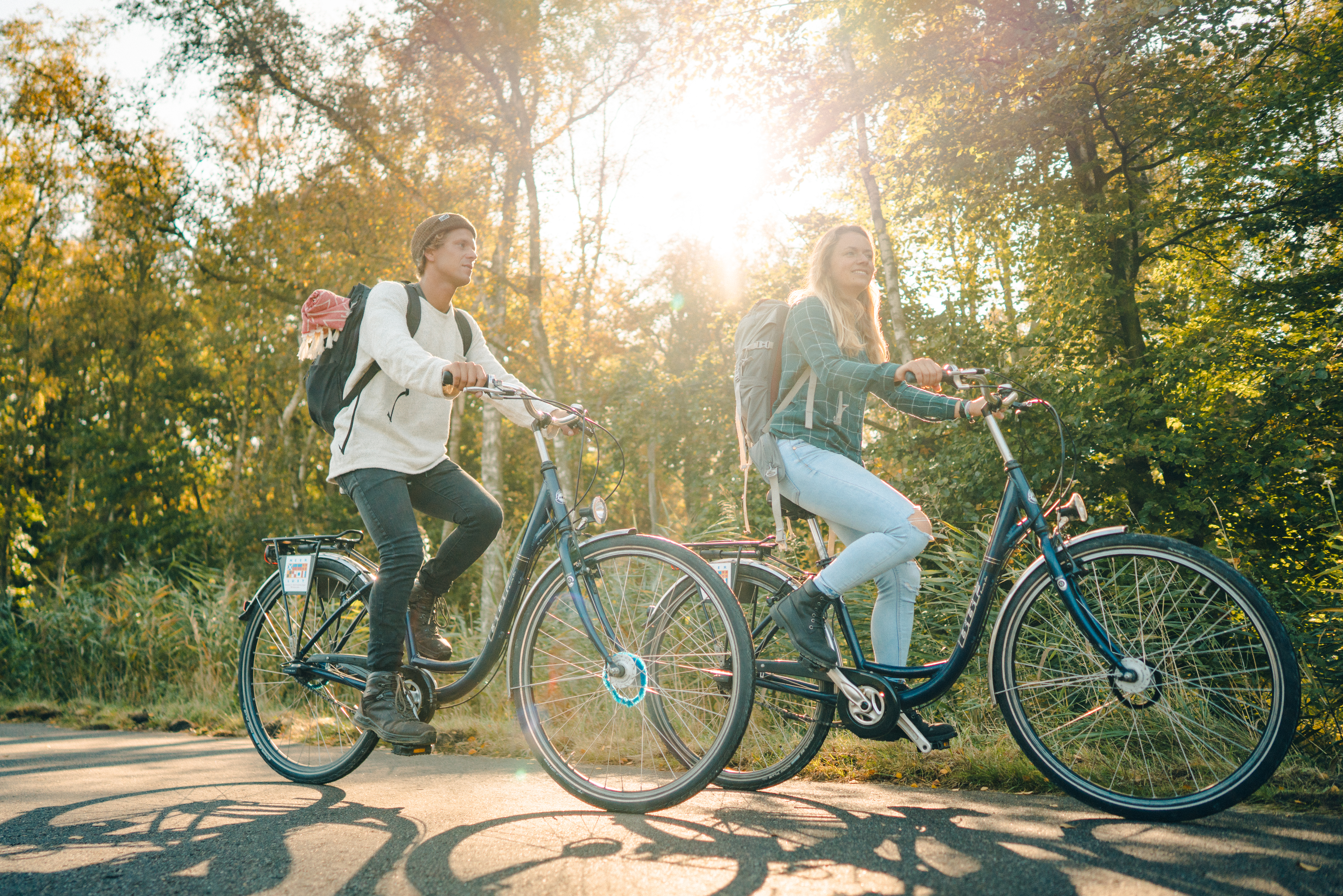 Ein Mann und eine Frau fahren auf Fahrrädern auf einem Asphaltweg durch den Wald. Das Bild wurde von unten aufgenommen, die Radfahrer sind im Fokus. Durch die beiden scheint hell die Sonne
